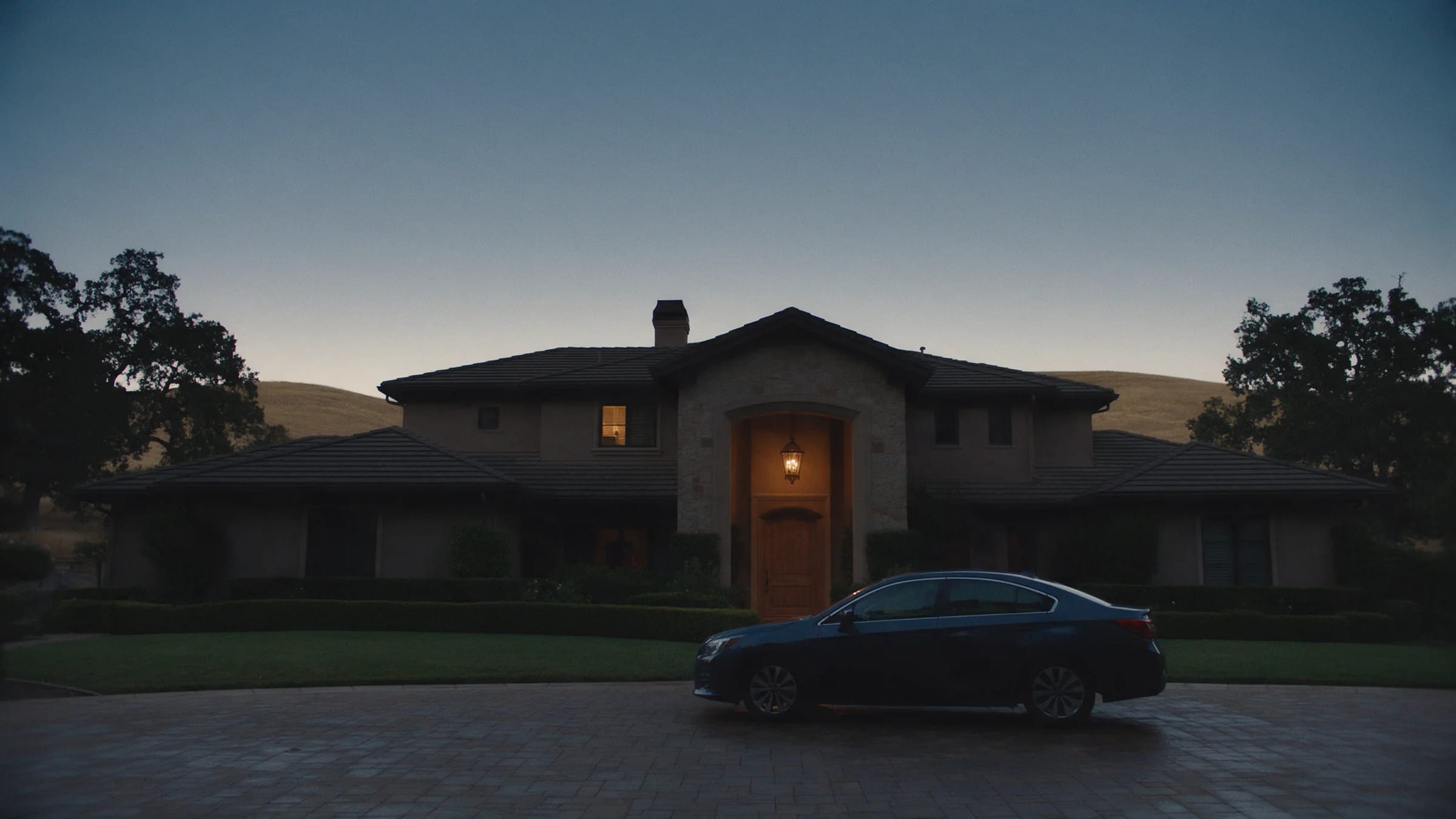 The front of a large Pleasanton house twenty minutes after sunset: a paver driveway, a lantern lit over the entry, the door shut, and the dry hills behind the roofline.